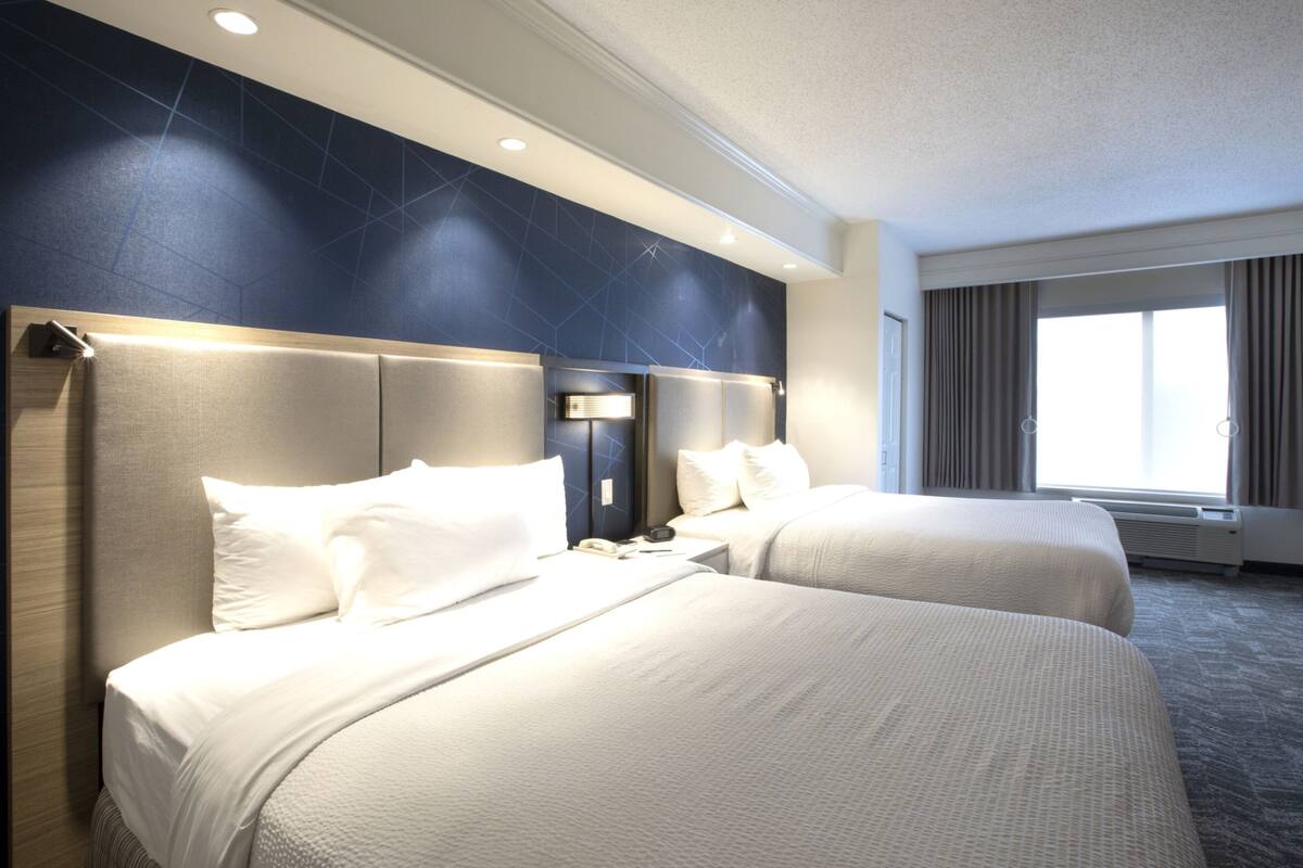A hotel room with two beds, a nightstand, a wall-mounted TV, and a large window with curtains.