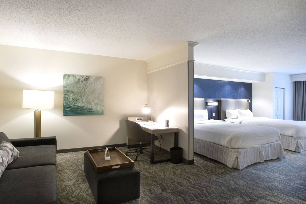 A hotel room with a bed, a desk, a chair, a lamp, a painting, and a trash can.