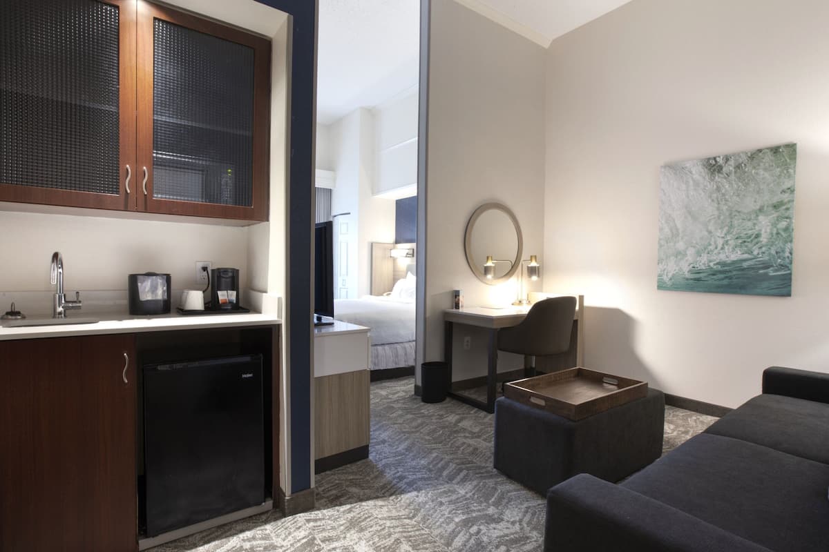 A hotel room with a kitchenette, a bed, a desk with a chair, a small sofa, and a painting on the wall.