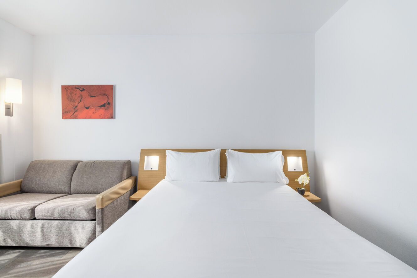 A hotel room with a bed, a sofa, two wall-mounted lamps, and a painting on the wall.