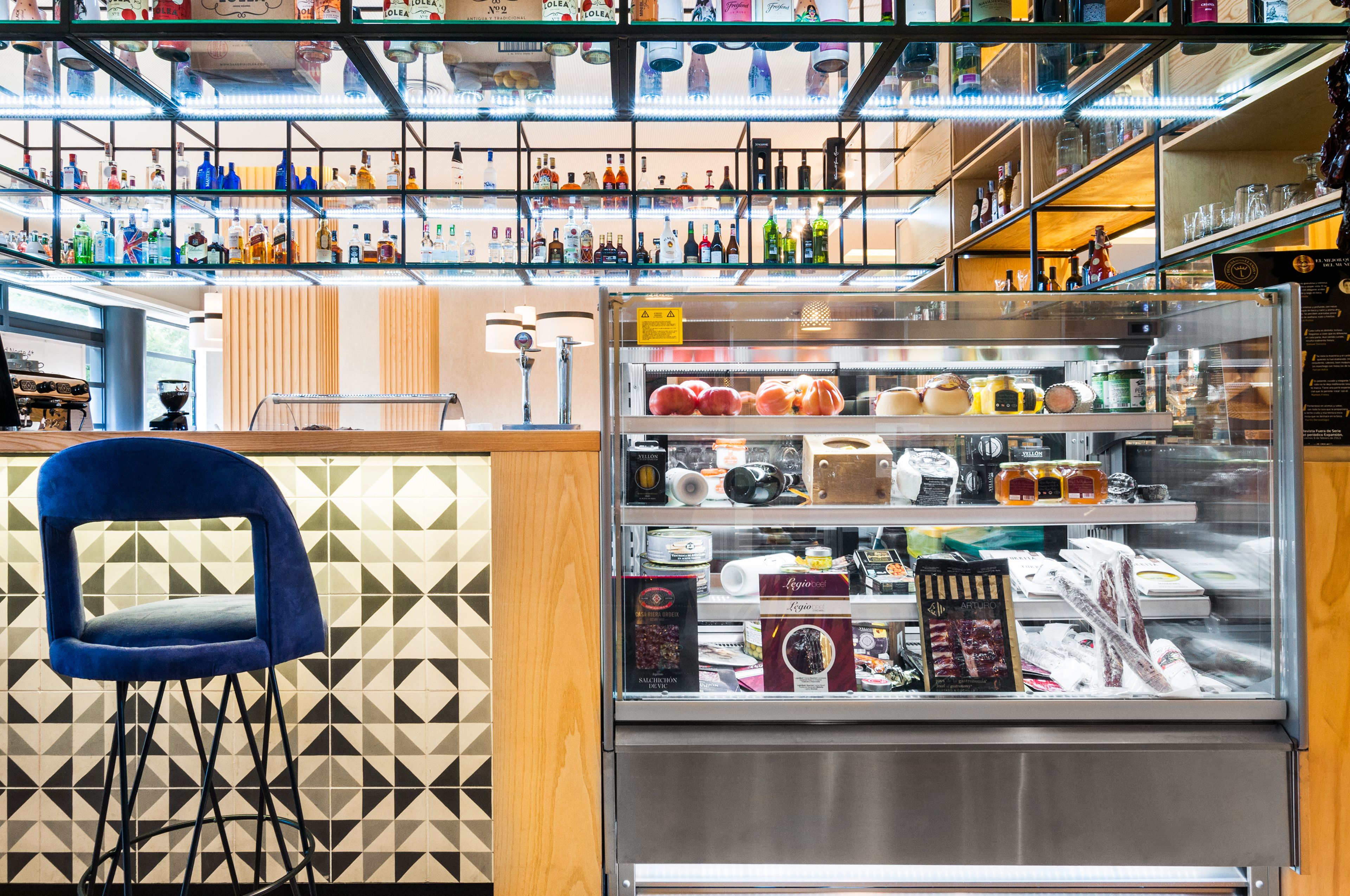 Una zona de bar con estantes llenos de botellas, una vitrina con alimentos y una silla azul.