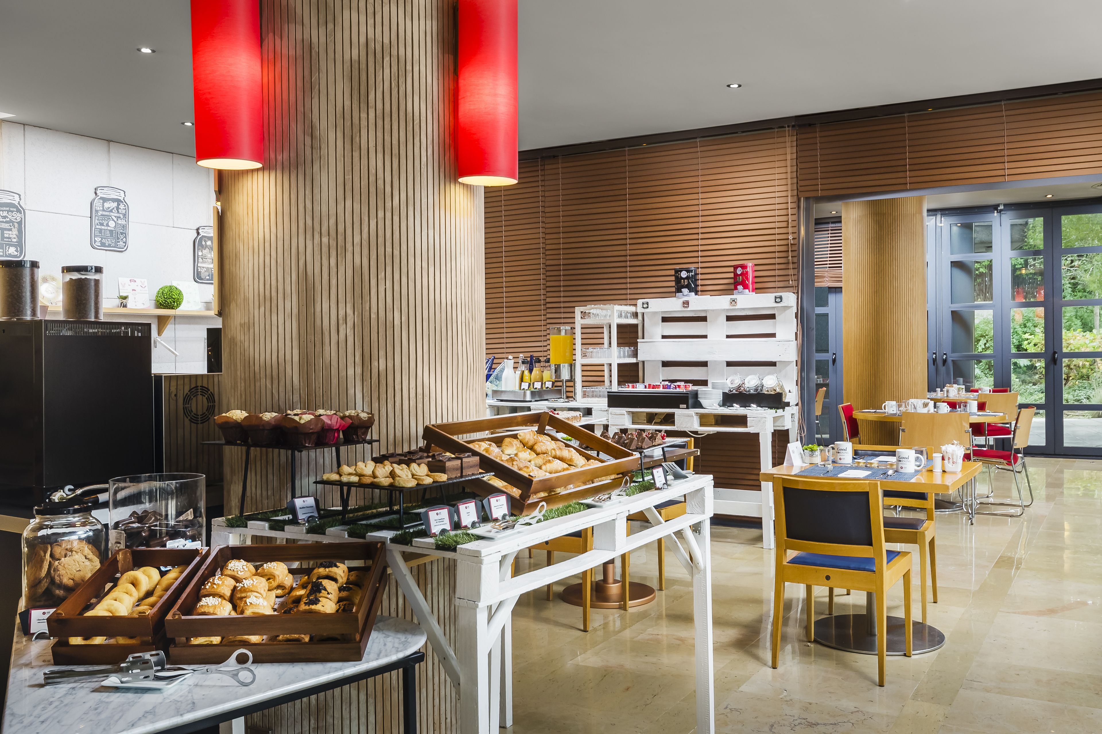 A modern breakfast area with a variety of pastries and beverages, wooden accents, and a large window offering a view of greenery.