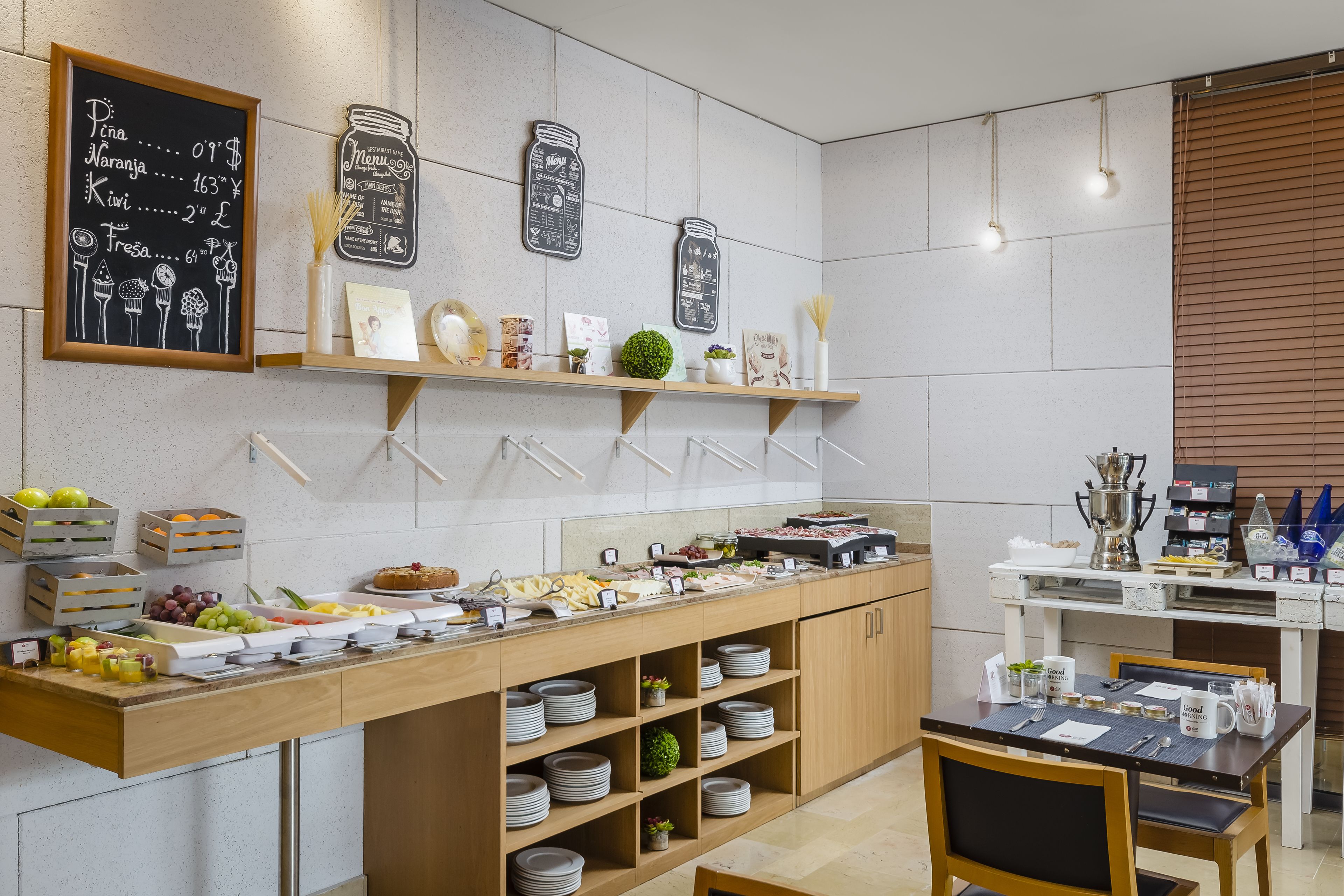 A modern kitchen with a buffet setup, including a chalkboard menu, shelves with kitchenware, and a dining area with a table set for two.