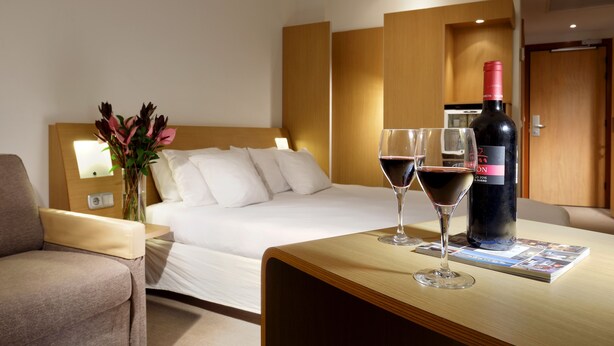 Une chambre d’hôtel avec un lit, une table de chevet, une bouteille de vin, deux verres à vin et un vase avec des fleurs.