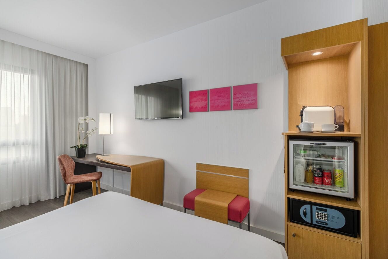 Een hotelkamer met een bed, een bureau, een stoel, een televisie, een koelkast en een raam met gordijnen.