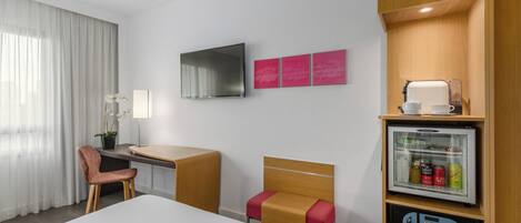 Habitación de hotel con cama, escritorio, silla, televisor, refrigerador y ventana con cortinas.