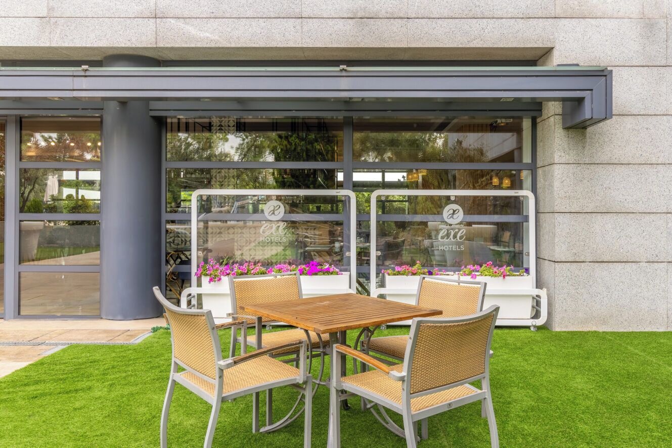 Un área de estar al aire libre con mesa y sillas, fachada de vidrio con el nombre del hotel y plantas en macetas.