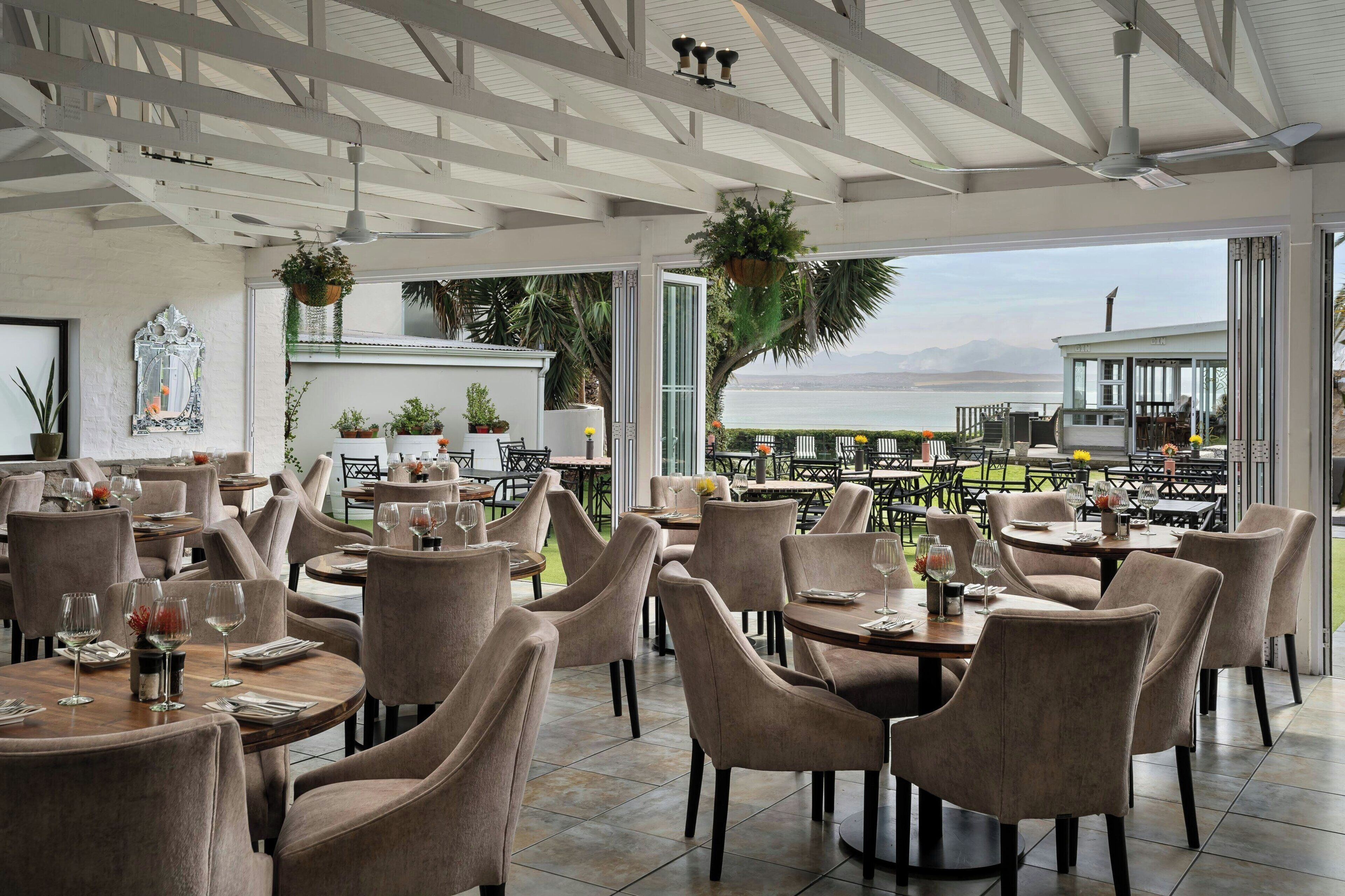 A restaurant with round tables and chairs, large windows, and hanging plants.