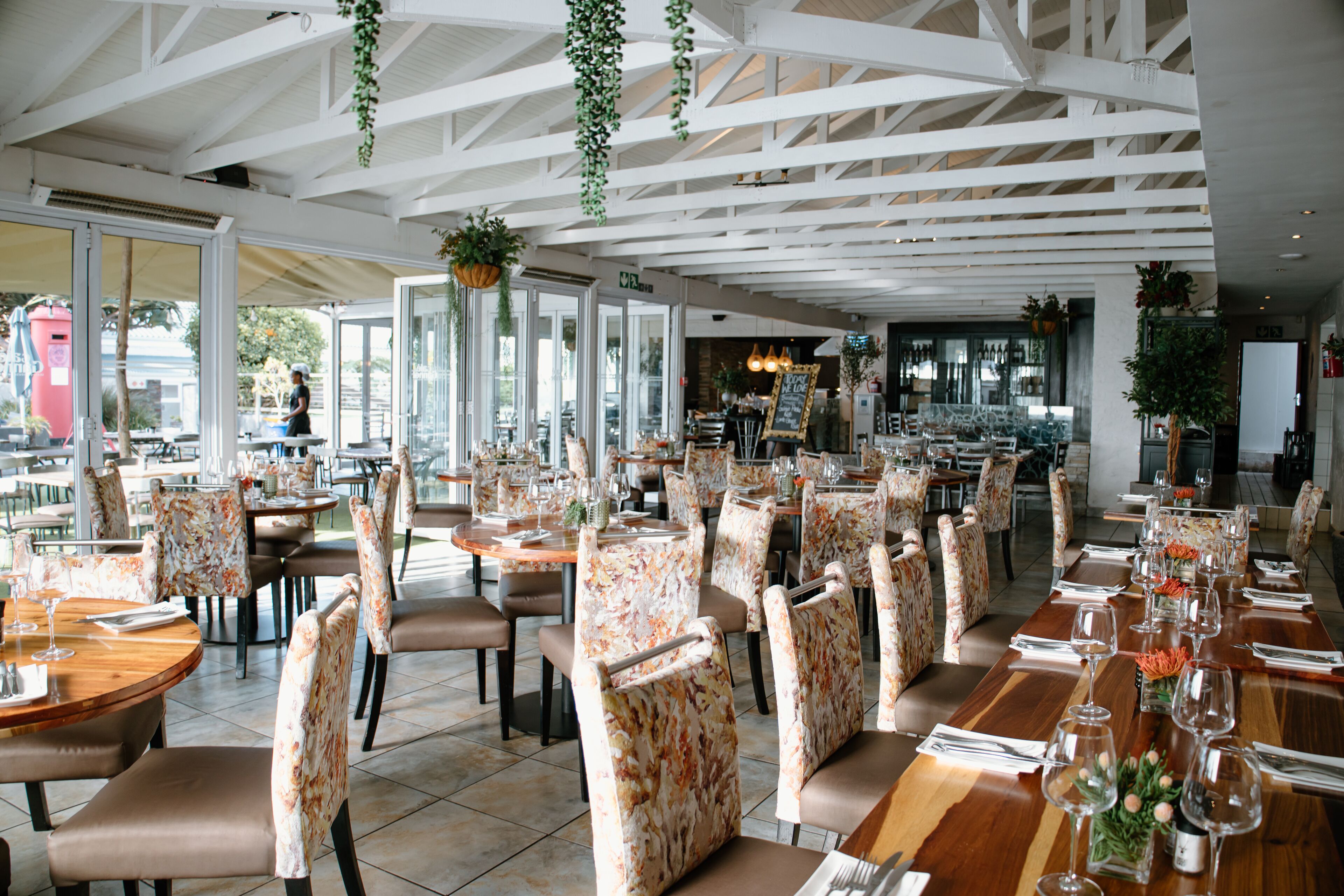 A spacious restaurant with high ceilings and hanging plants, featuring numerous tables and chairs arranged for dining.