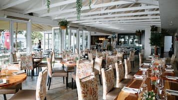 A spacious restaurant with high ceilings and hanging plants, featuring numerous tables and chairs arranged for dining.