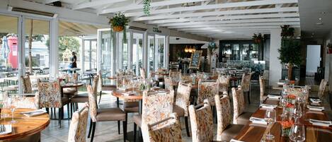 A spacious restaurant with high ceilings and hanging plants, featuring numerous tables and chairs arranged for dining.