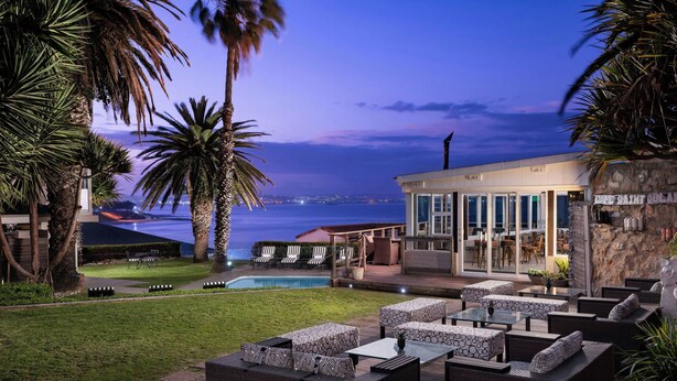 An outdoor patio with seating, a pool, and a view of the ocean at dusk. - Protea Hotel by Marriott Mossel Bay (Mossel Bay)