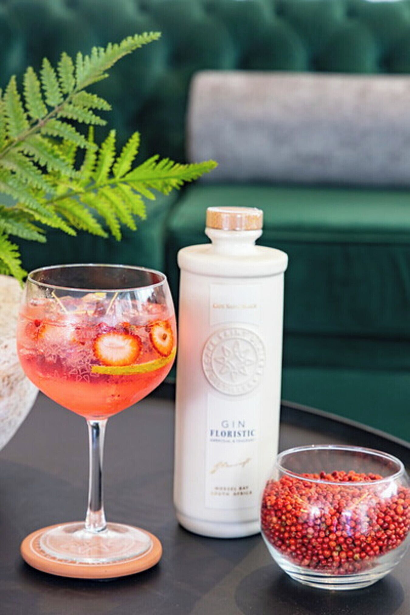 Een cocktailglas met een roze drankje, een fles gin en een schaal rode bessen op een tafel.