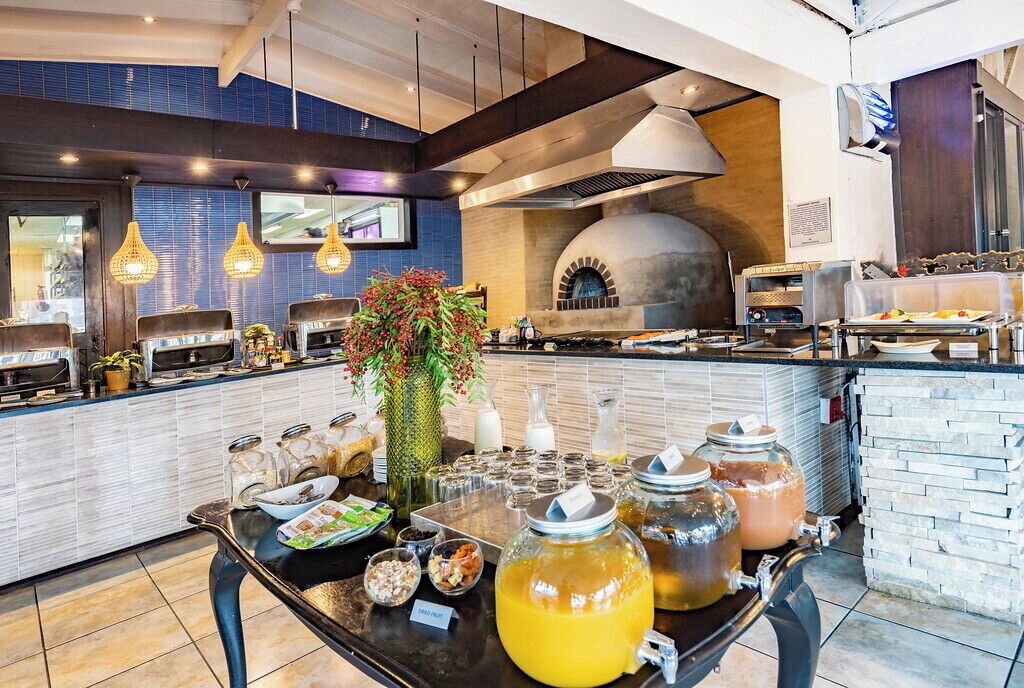 Een buffetopstelling met diverse gerechten en dranken, een grote vaas met bloemen, en een pizzaoven op de achtergrond.