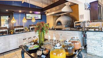 Een buffetopstelling met diverse gerechten en dranken, een grote vaas met bloemen, en een pizzaoven op de achtergrond.