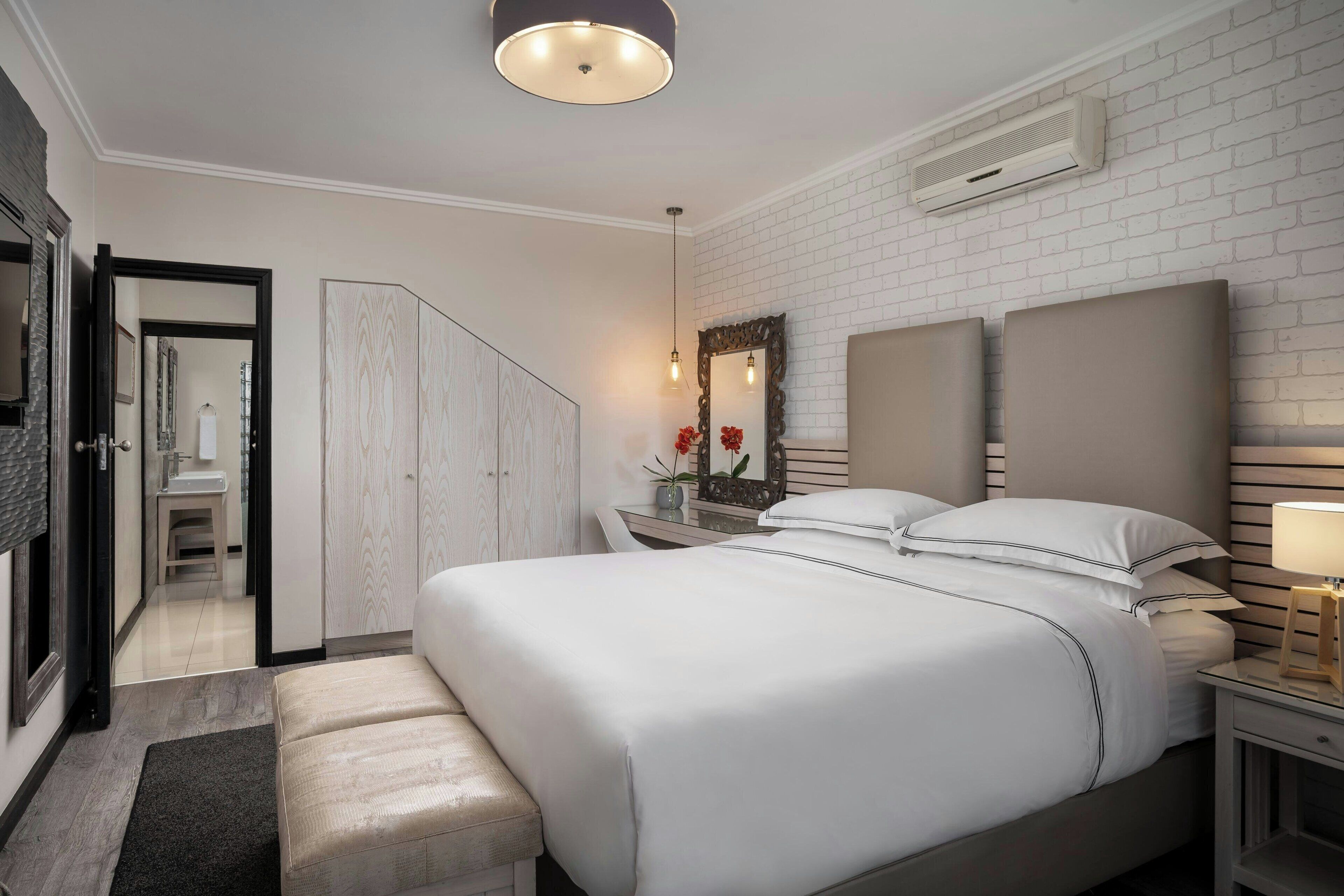 A modern hotel room with a large bed, a bedside table with a lamp, a mirror, and a wardrobe.