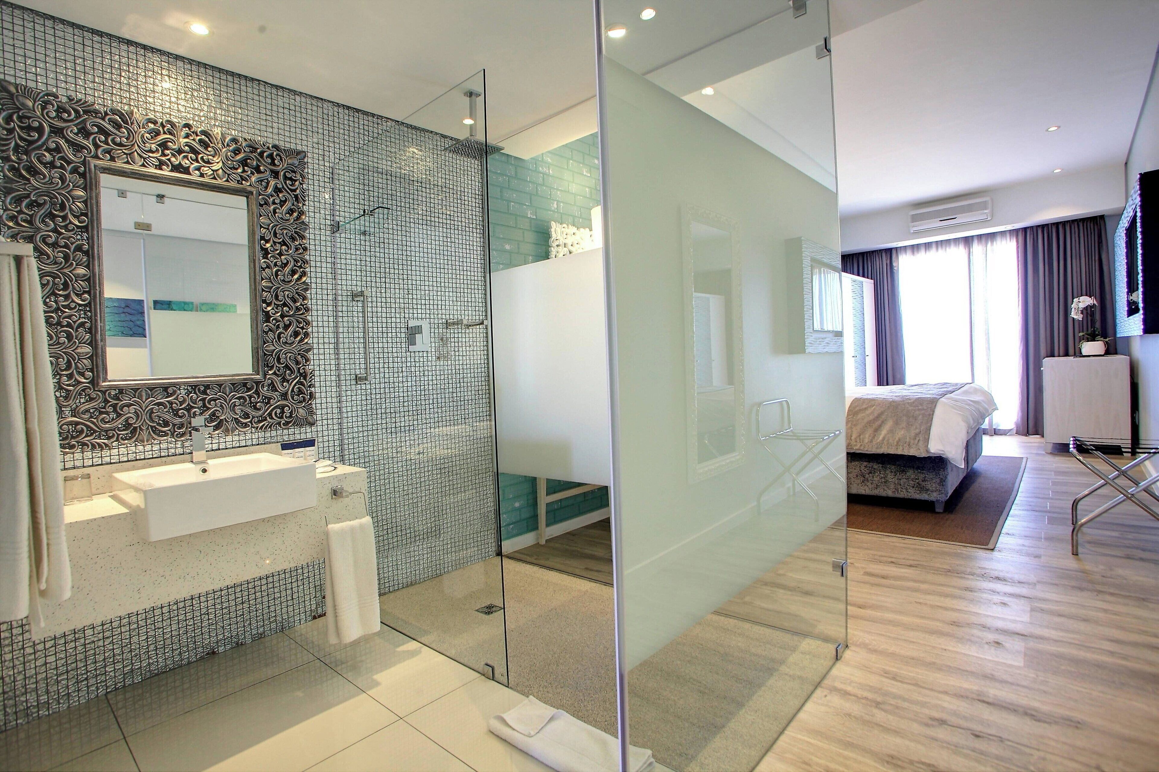 Een moderne badkamer met een douchecabine van glas, een wastafel met spiegel, en een bed in de aangrenzende kamer.