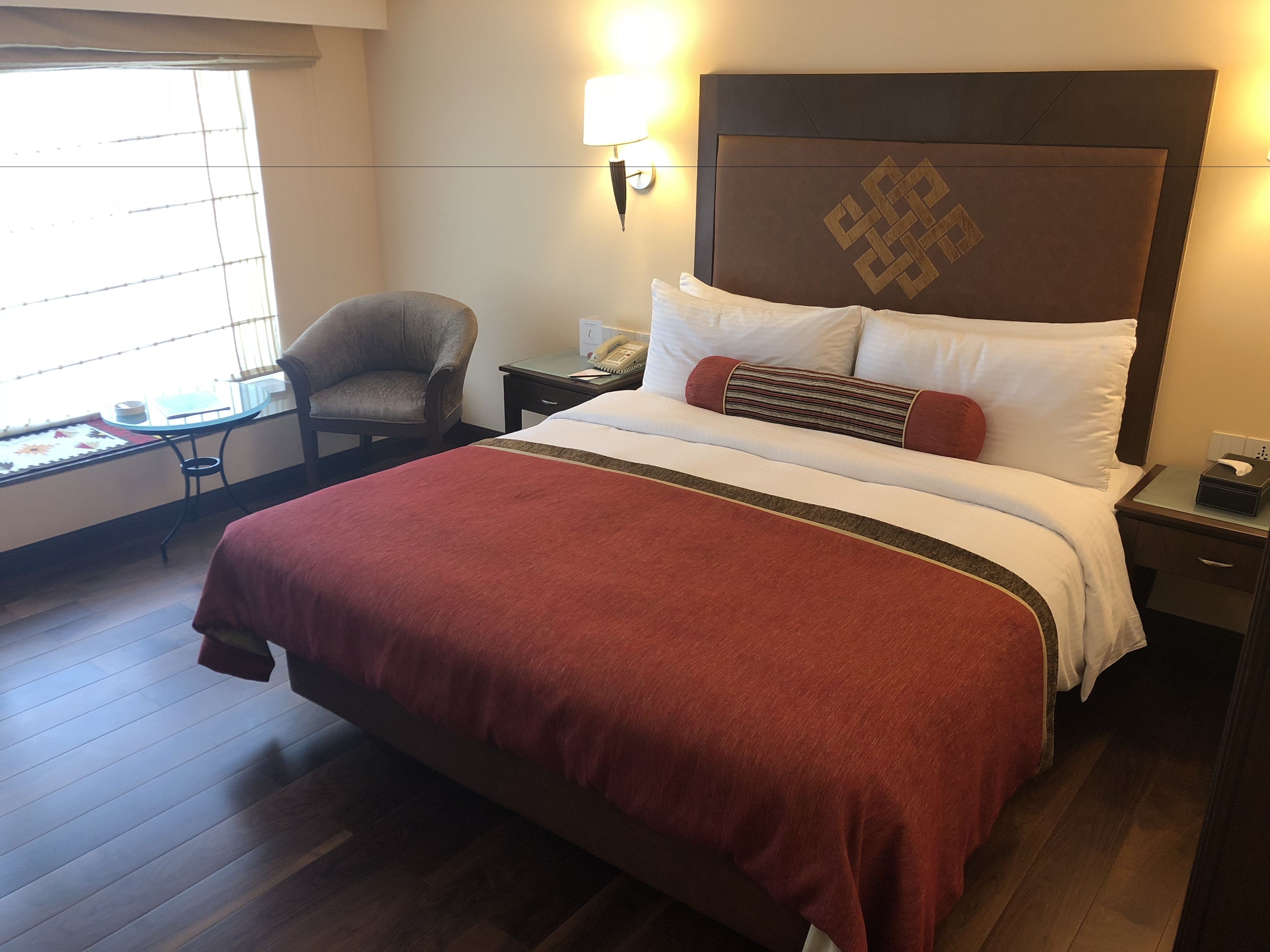 A hotel room with a large bed, a chair, a bedside table with a lamp, and a window with blinds.