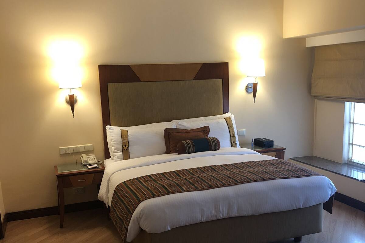 Quarto de hotel com uma cama grande, duas mesas de cabeceira, um criado-mudo, telefone e luminárias fixadas na parede.