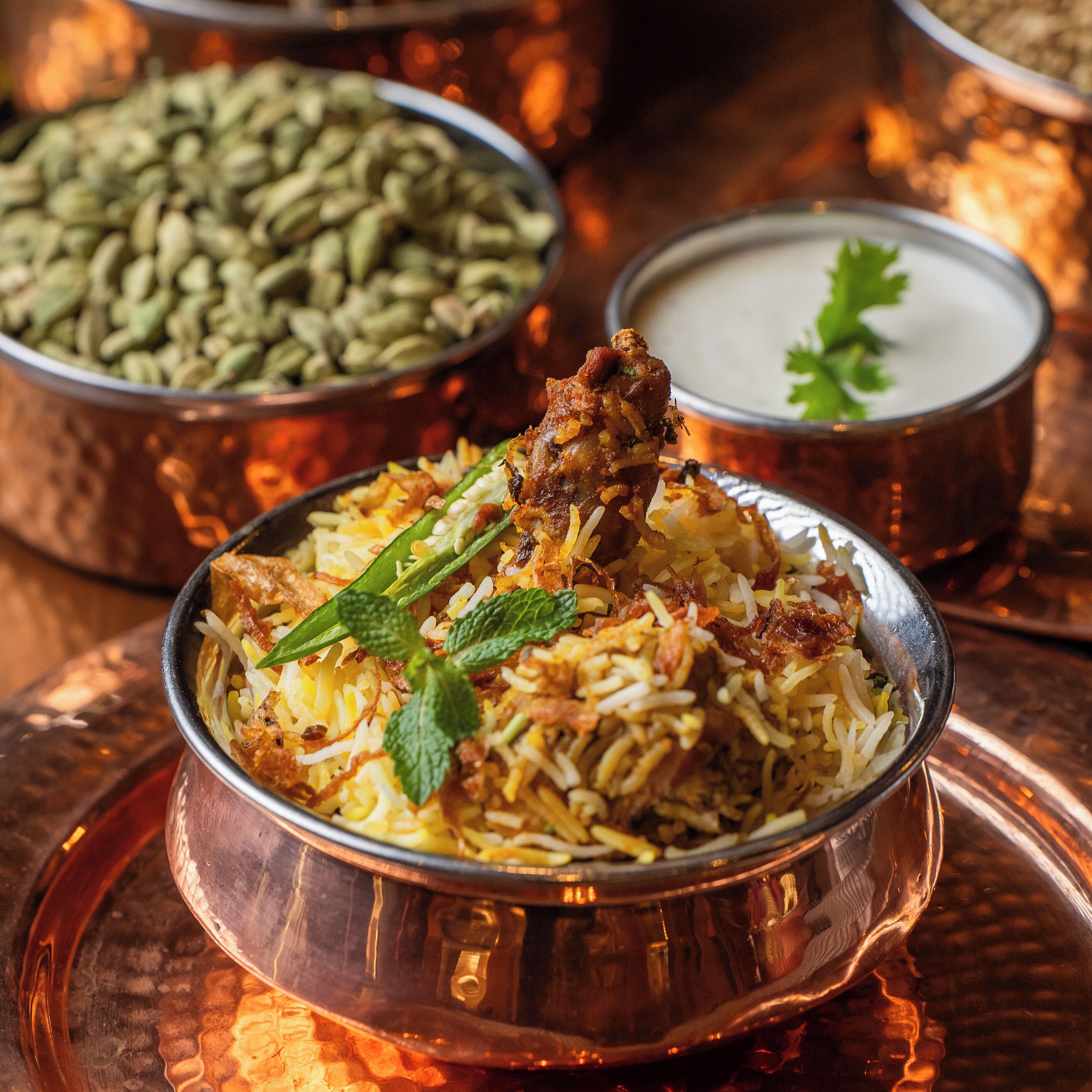 A bowl of biryani with a piece of meat on top, garnished with mint leaves, and surrounded by two small containers, one with a green substance and the other with a white substance.