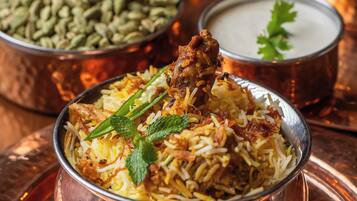 A bowl of biryani with a piece of meat on top, garnished with mint leaves, and surrounded by two small containers, one with a green substance and the other with a white substance.