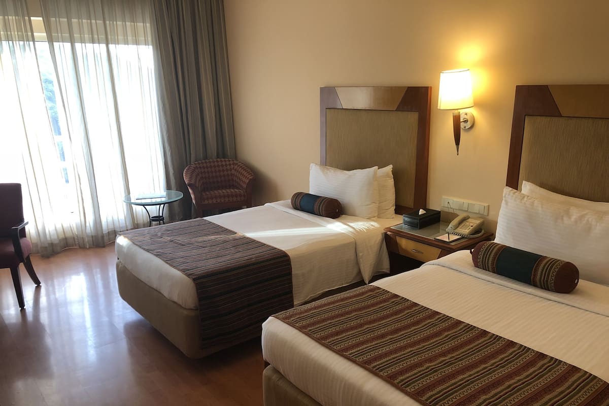 Quarto de hotel com duas camas, uma cadeira, uma mesinha e um abajur.