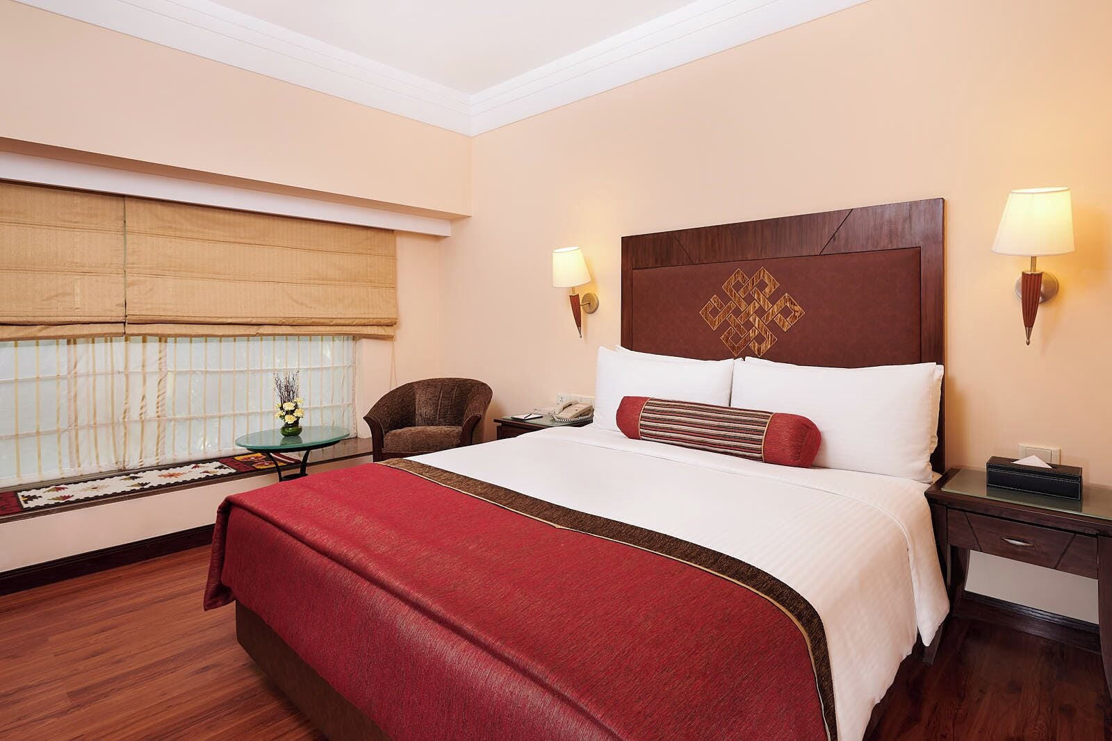 A hotel room with a large bed, a chair, a small table with a vase, and a window with blinds.