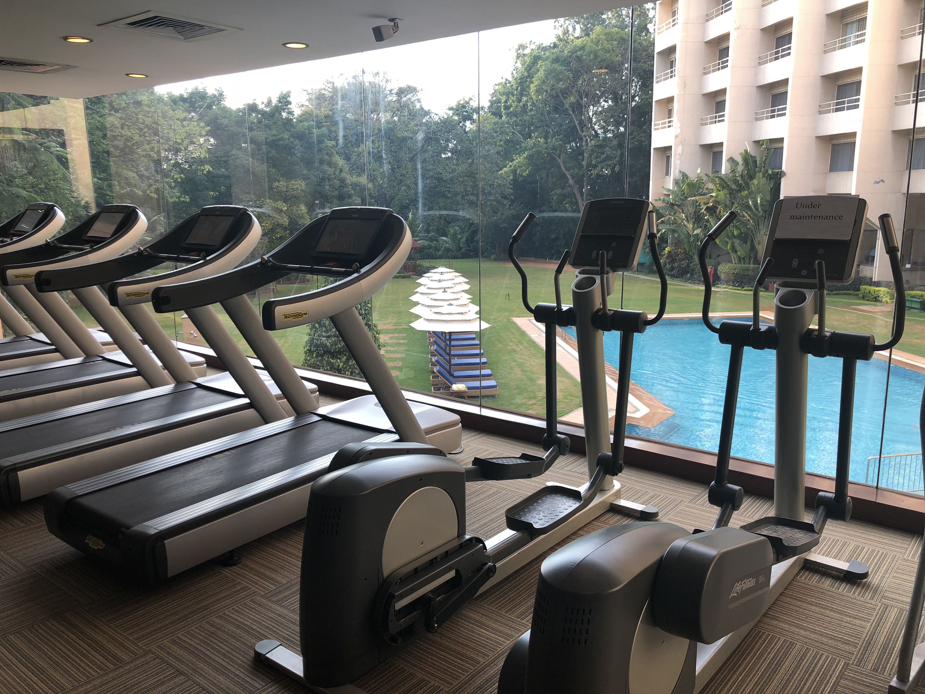A gym with treadmills and elliptical machines, overlooking a swimming pool and lush greenery.