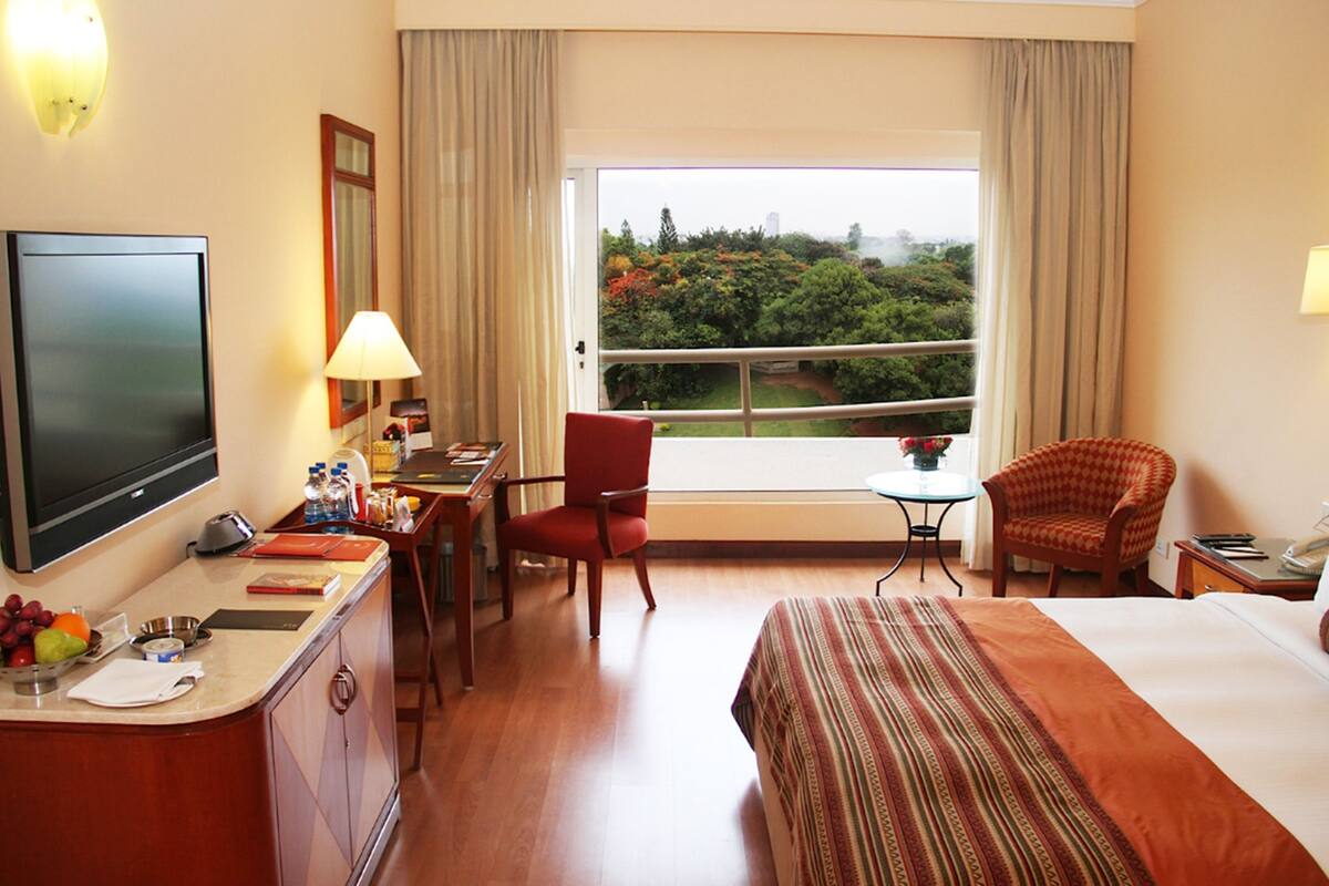 Quarto de hotel com uma cama, uma escrivaninha, uma televisão, uma cadeira, uma mesa e vista para o verde lá fora.