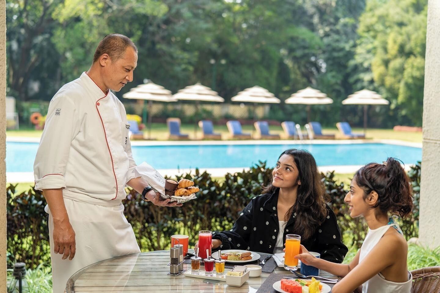 A dining table with food and drinks, a pool in the background, and lounge chairs.