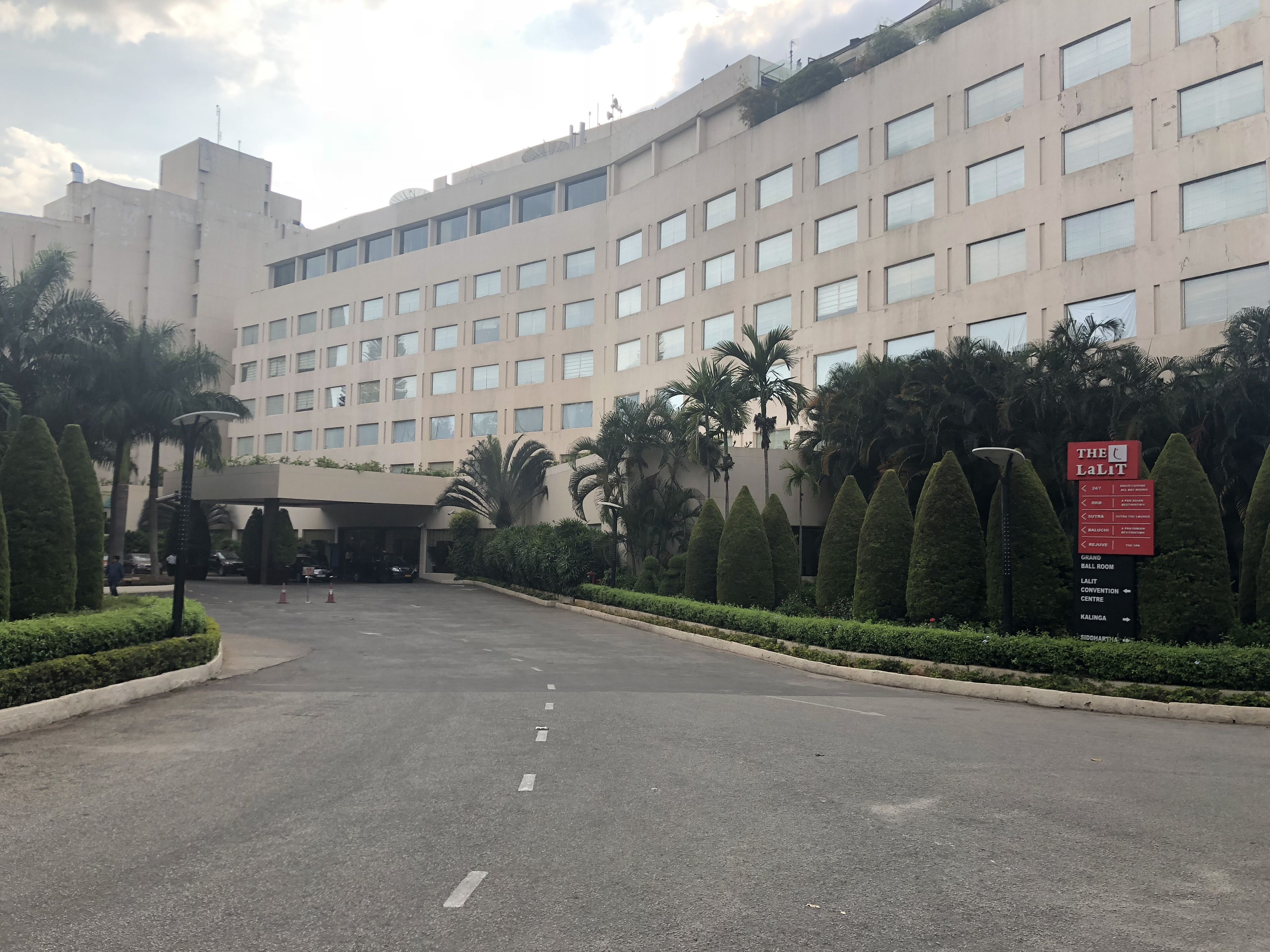 A large hotel with a central entrance, surrounded by well-maintained hedges and palm trees.