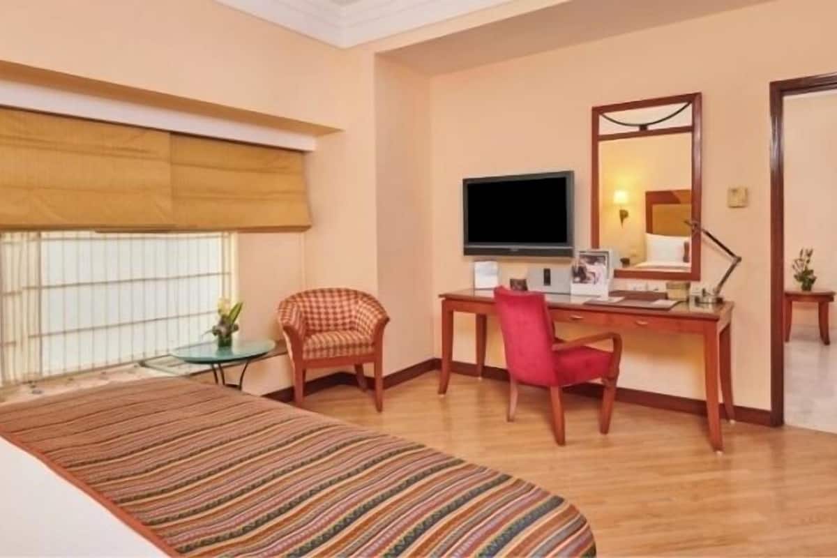 Quarto de hotel com cama, escrivaninha com cadeira, televisão e espelho.