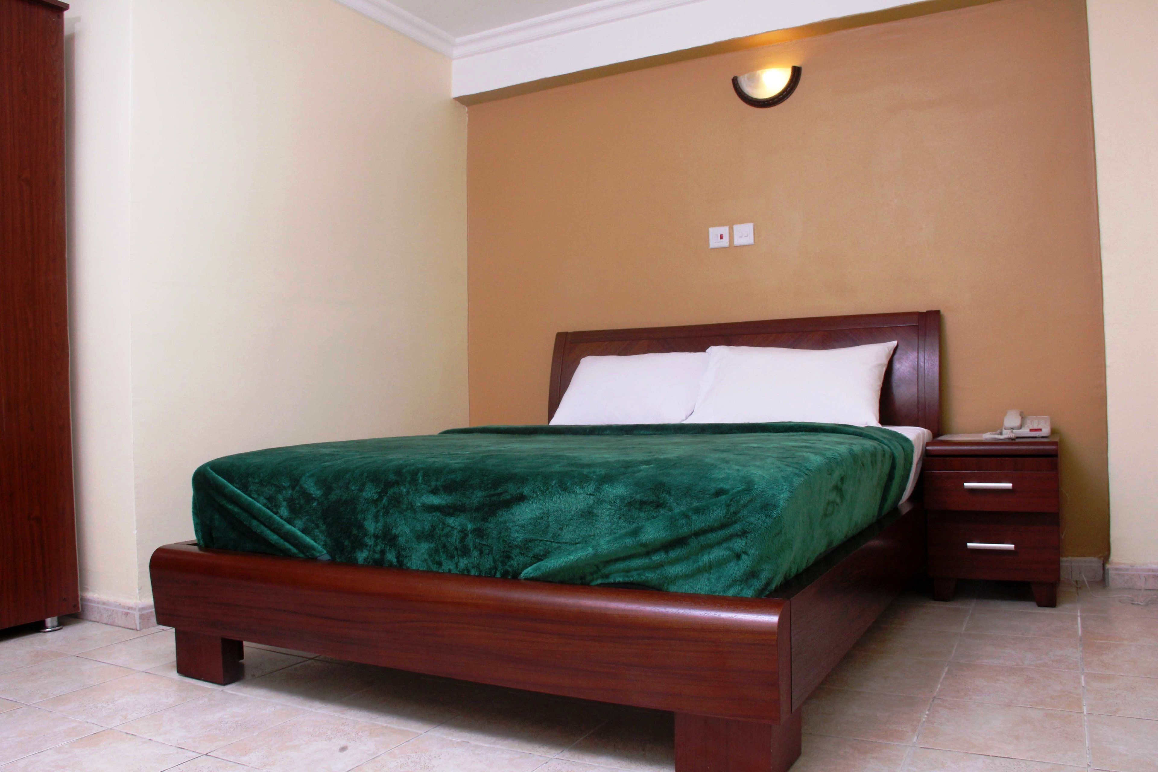 Una cama bien hecha con colcha verde y almohadas blancas, una mesita de noche de madera con un teléfono, y un aplique de pared.