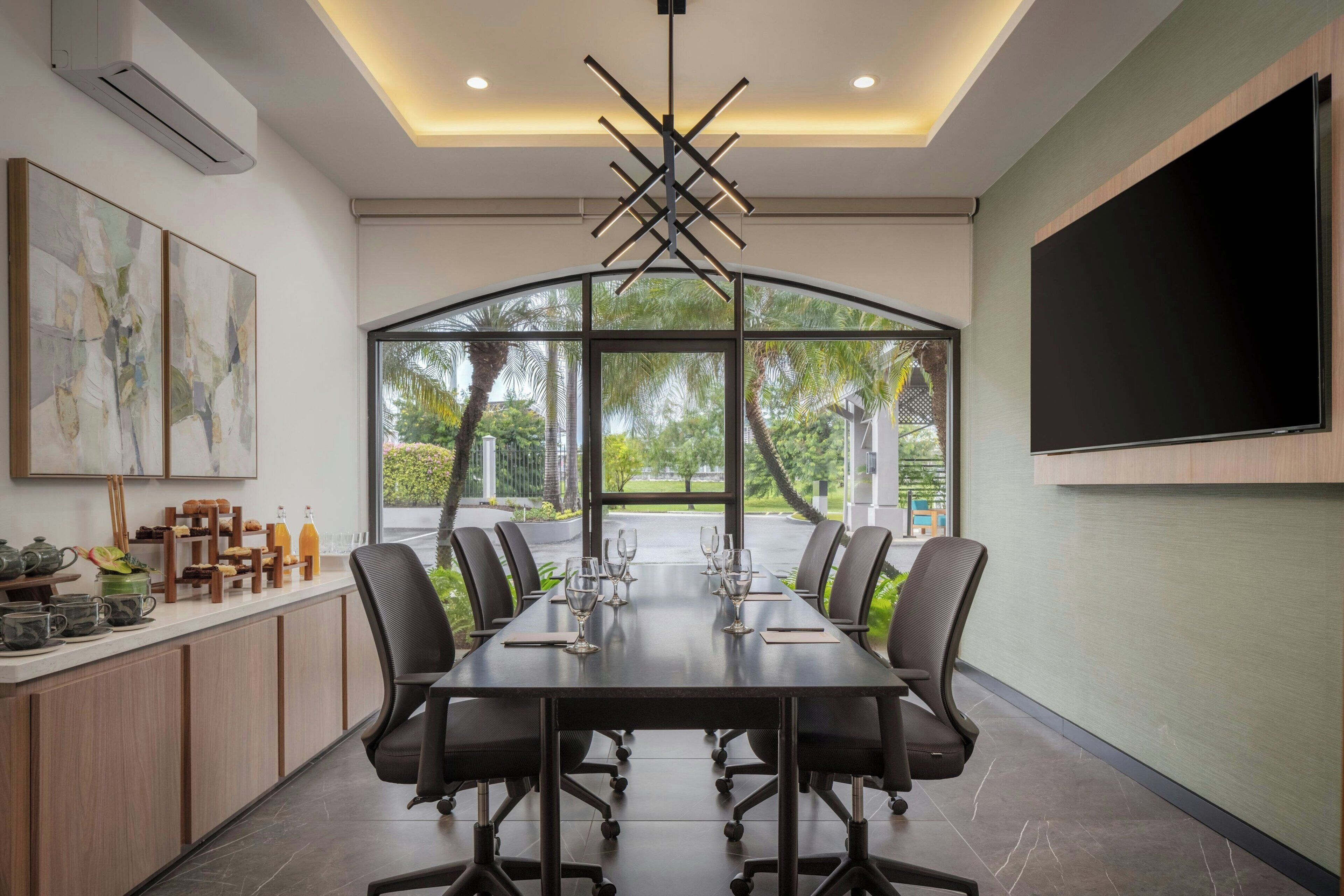 Un salón de conferencias con una mesa larga, sillas, un televisor y vistas al verdor exterior.