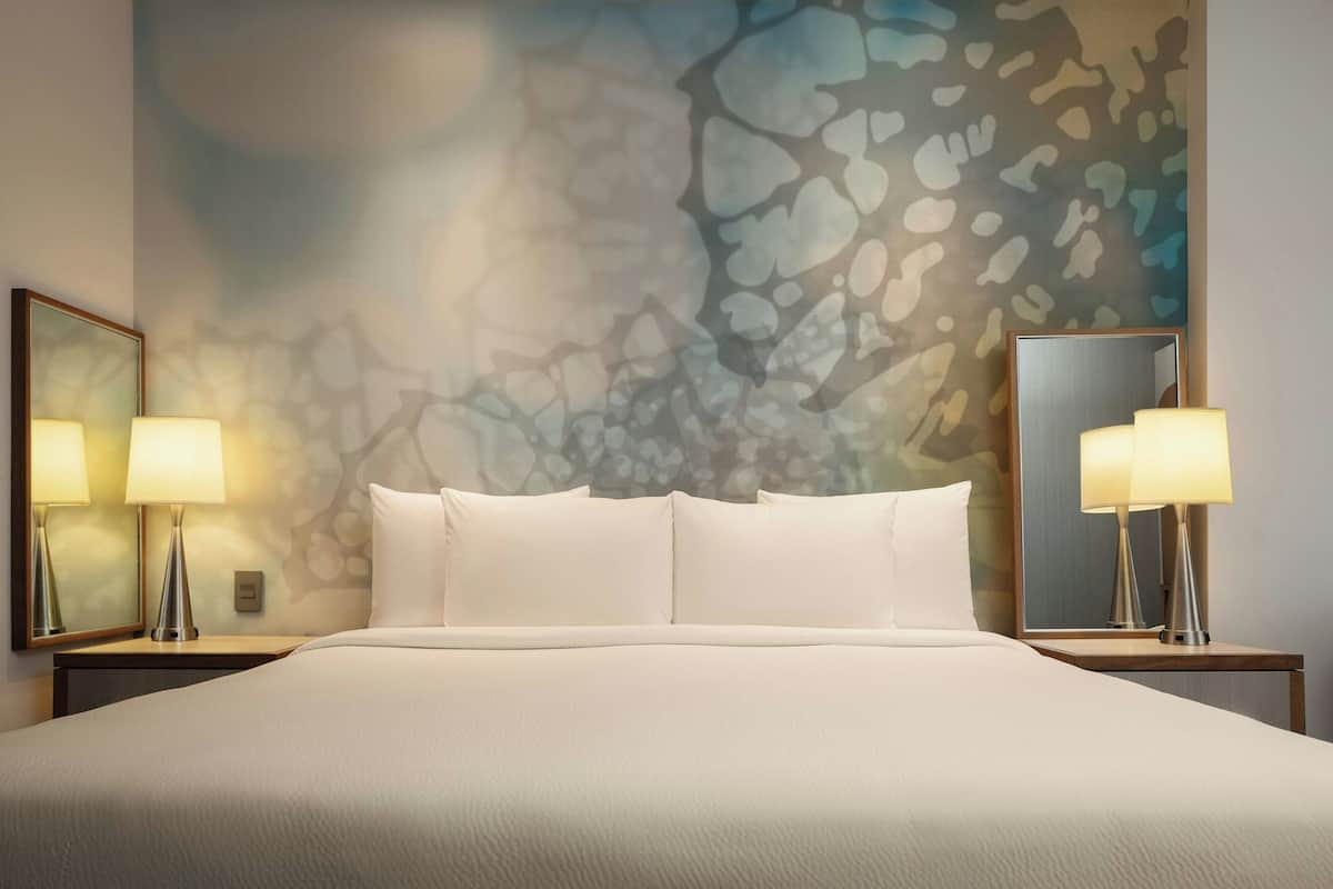 Una cama bien tendida con sábanas blancas, dos lámparas de noche y un cuadro abstracto grande en la pared.