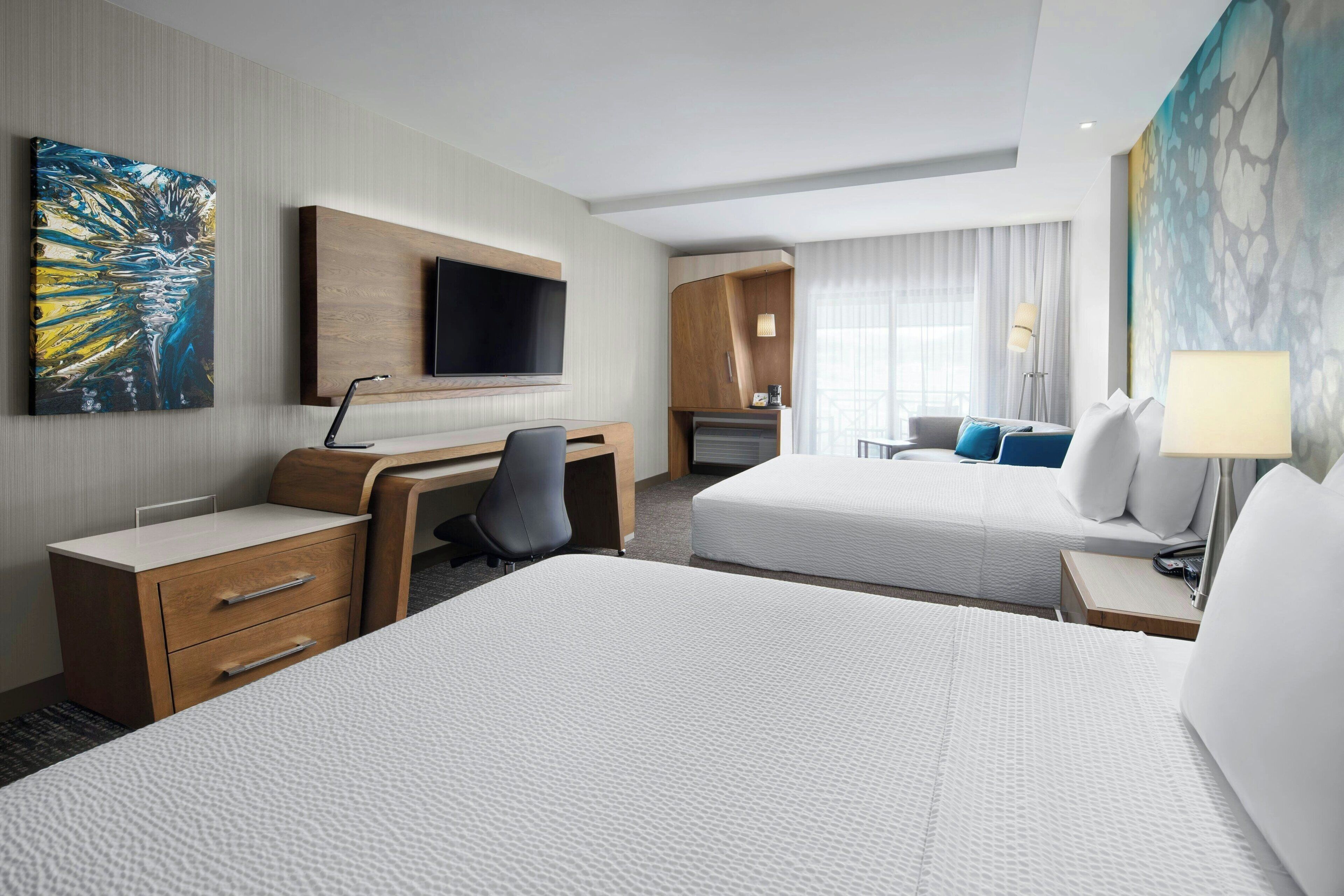 A hotel room with two beds, a desk, a TV, and artwork on the walls.