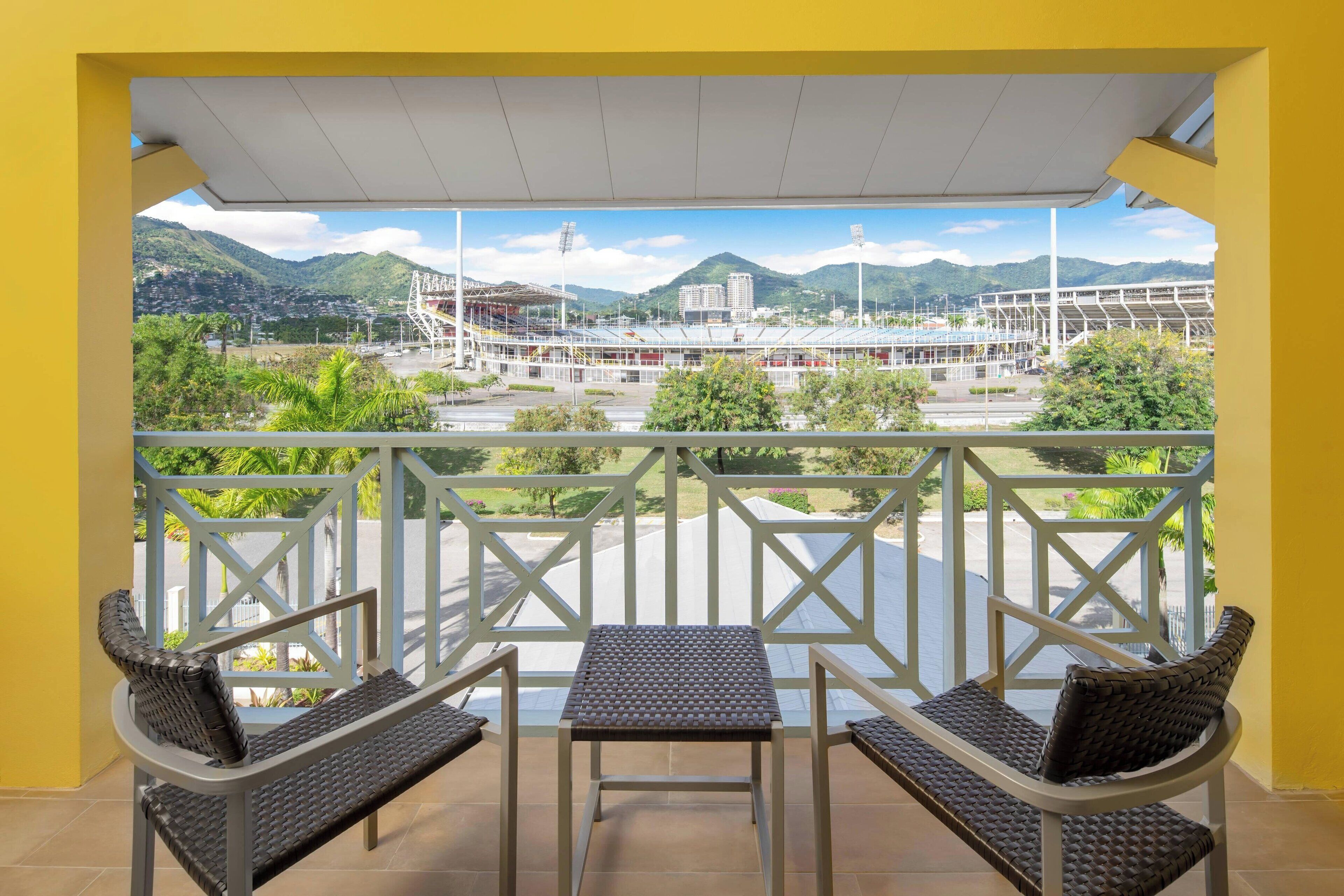 Uma varanda com duas cadeiras de vime e uma mesa pequena, com vista para um estádio e montanhas.