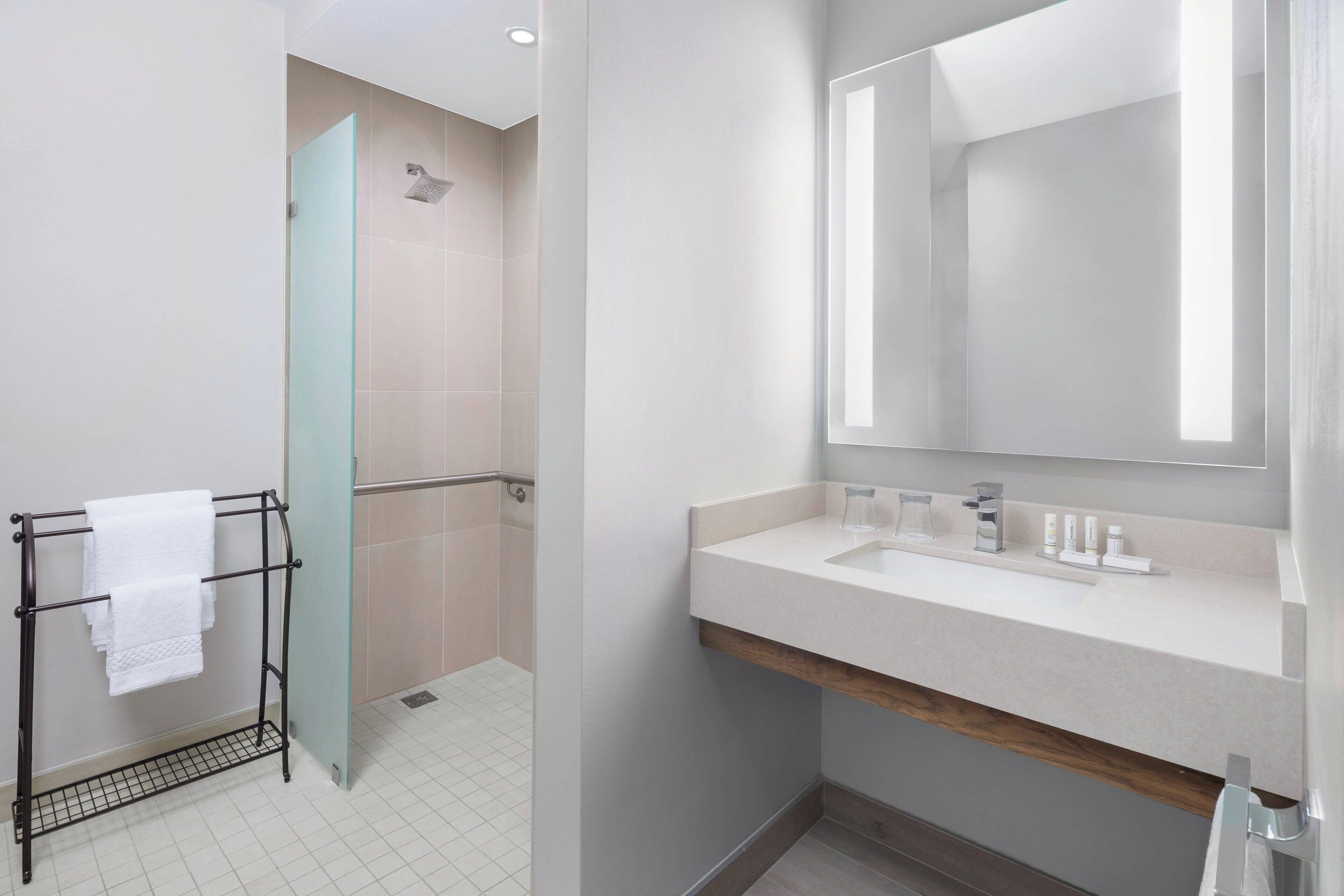 A modern bathroom with a glass shower enclosure, a towel rack with white towels, a large mirror, and a wooden vanity.