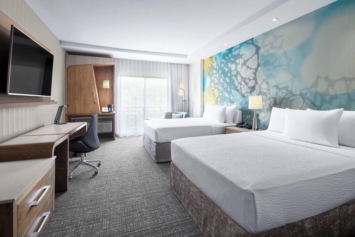 Habitación de hotel con una cama grande, un escritorio con silla, un televisor en la pared y un amplio ventanal con cortinas.