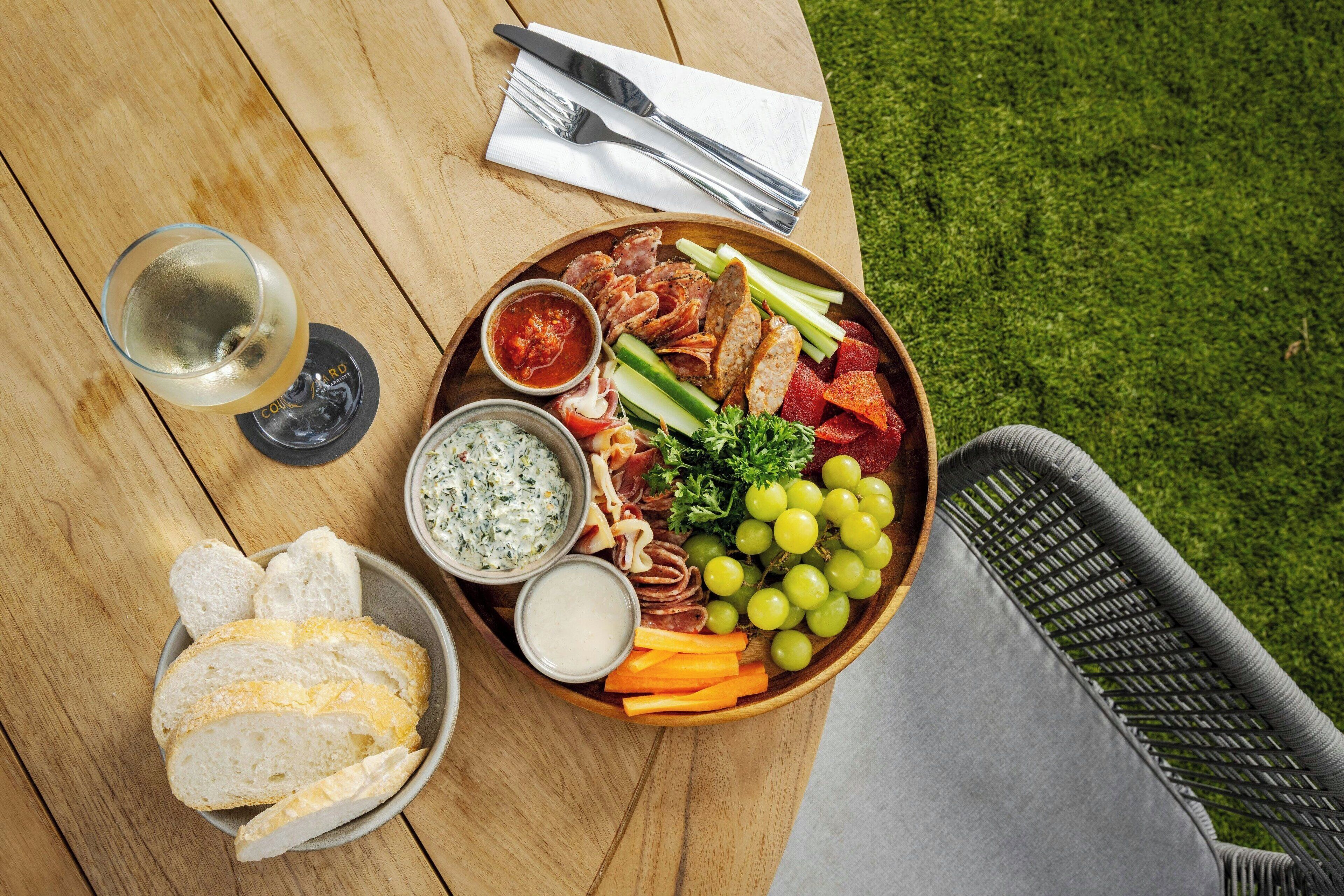 A wooden table with a charcuterie board, a glass of white wine, and a wicker chair.