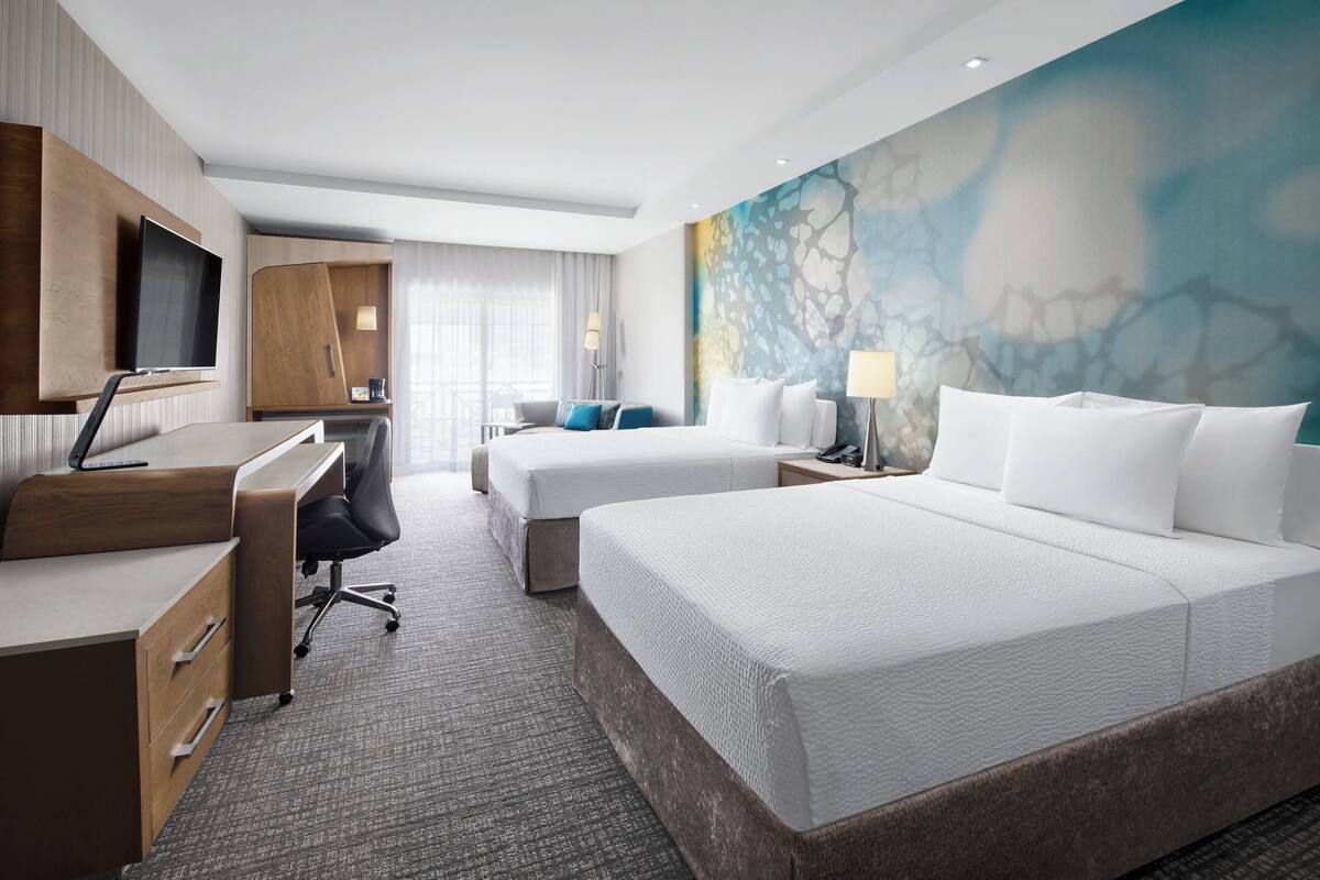 Habitación de hotel con dos camas, un escritorio, un televisor y un mural abstracto grande en la pared.