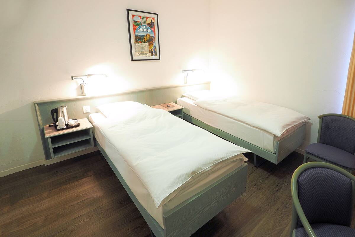 A hotel room with two single beds, a nightstand with a coffee maker, a chair, and a framed picture on the wall.