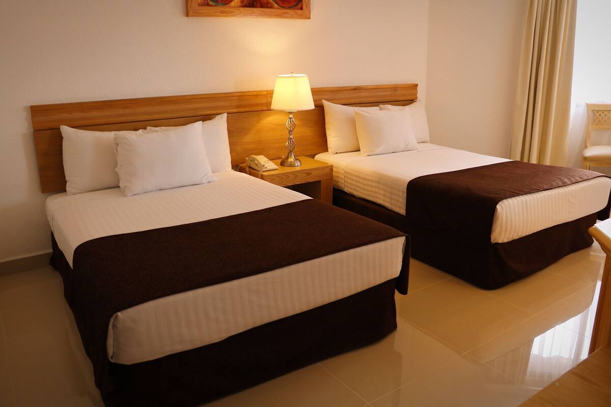 Dos camas individuales con cabeceros de madera, una mesita de noche con lámpara y una silla en una habitación de hotel.