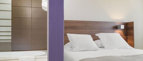 A modern hotel room with a wooden headboard, white bedding, and a bathroom with a bathtub and towel rack.