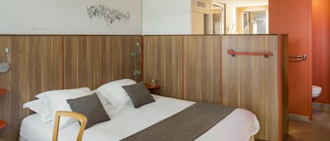 A modern hotel room with a large bed, a desk with a laptop, a wooden headboard, and a wardrobe.