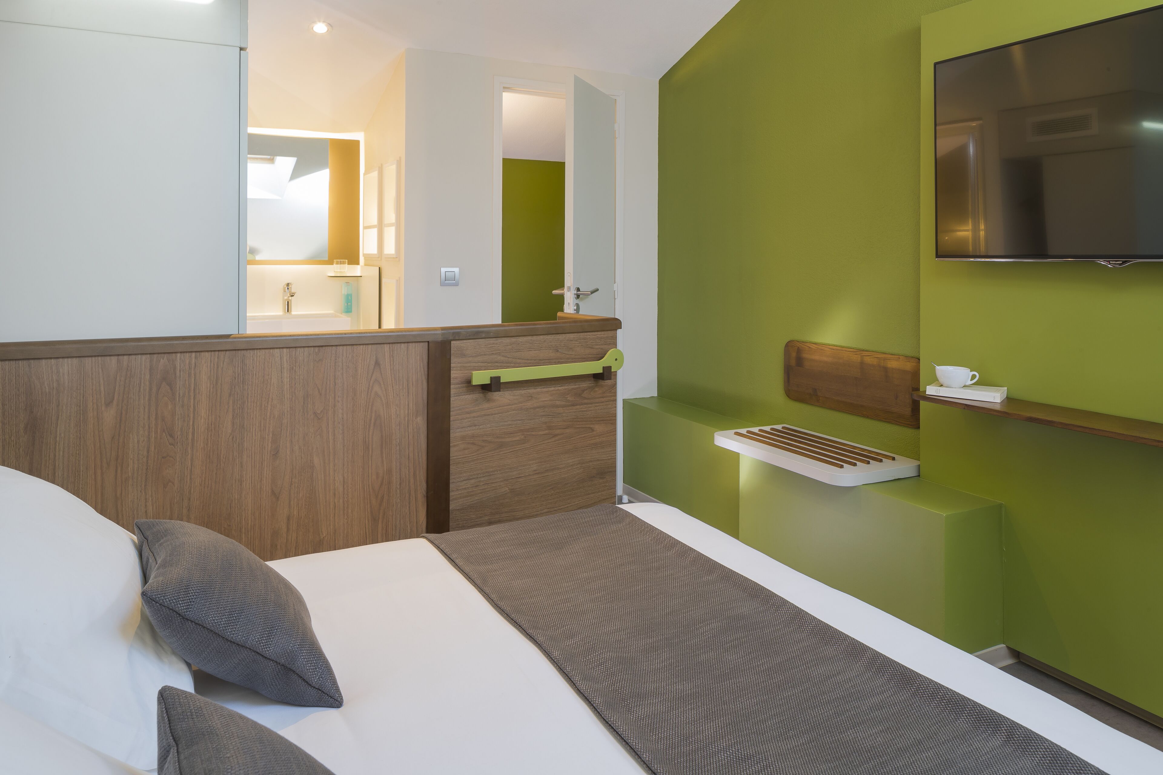 A modern hotel room with a bed, a wooden headboard, a green accent wall, a mounted TV, and a small bedside table with a cup.