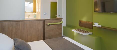 A modern hotel room with a bed, a wooden headboard, a green accent wall, a mounted TV, and a small bedside table with a cup.