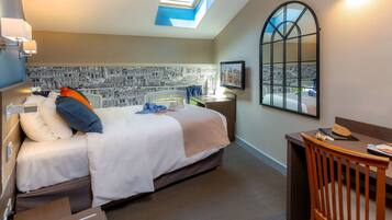 A hotel room with a bed, a desk, a chair, a desk lamp, a nightstand, a mirror, and a window with a view.