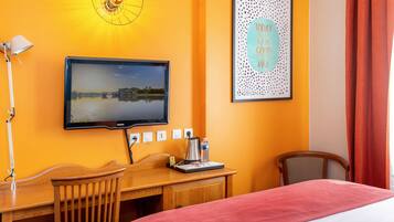 Habitación de hotel con cama, escritorio y silla, televisor, lámpara de pared, una cita motivacional en un marco y una ventana con cortinas.