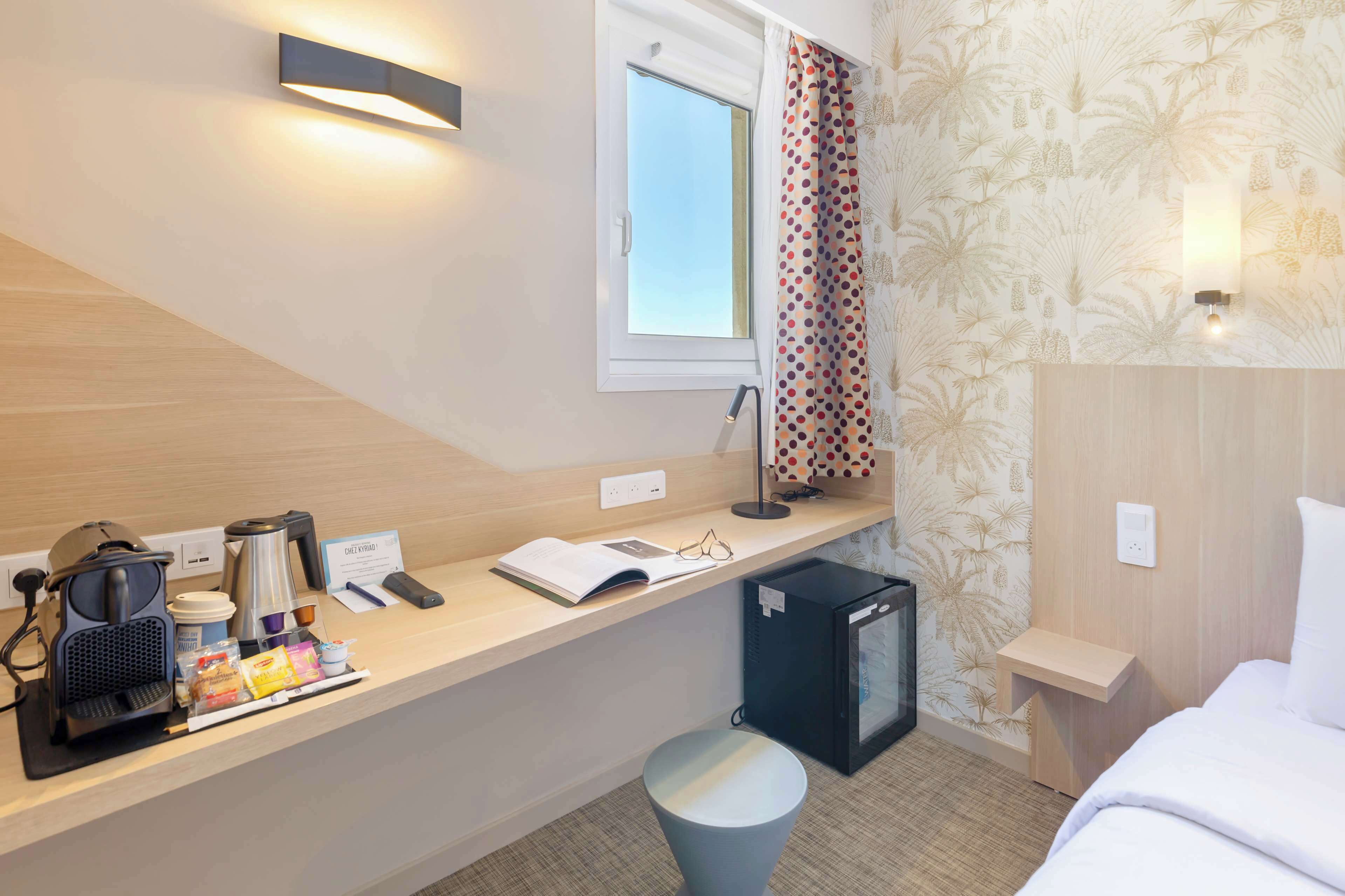 A hotel room with a wooden desk, a coffee machine, a mini fridge, a bed, and a window with curtains.